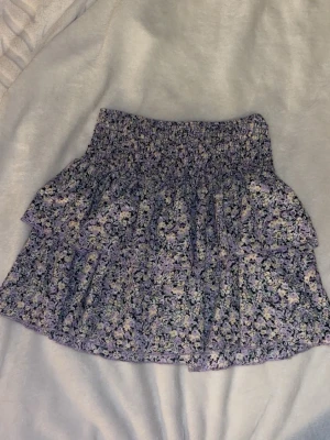 Lila blommig volangkjol - Supersöt kort kjol med blommigt mönster i lila, svart och gult. Kjolen har volanger och resår i midjan för en bekväm passform. Perfekt för dig som gillar en feminin och trendig stil.