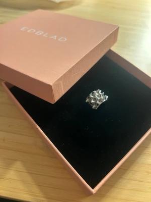 Silvrig ring från Edblad - Säljer en snygg silvrig ring från Edblad med cool pyramidformad design. Ringen har en bred form och är tillverkad i blankt metallmaterial som ger en modern och edgy look. Perfekt för dig som gillar statement-smycken. Säljer pga för liten, storlek 16