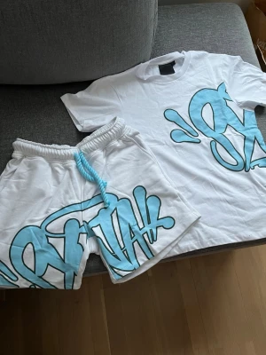 Syna World set - Säljer ett vitt Syna World set med t-shirt och shorts, båda med stort blå Syna tryck. Perfekt för dig som gillar streetwear och vill sticka ut med en unik look. Passar inte mig och är helt oanvända.