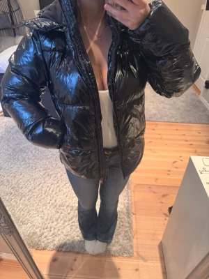 Svart glansig pufferjacka med huva från Nelly  - Säljer en svart glansig pufferjacka med huva och dragkedja framtill. Jackan har en oversized passform och quiltad design som ger en riktigt trendig look. Perfekt för kalla dagar när du vill vara både varm och snygg. Storleken på jackan är ”M” men jag som är en S & XS i vanliga fall tycker att den sitter väldigt fint på men fortfarande lite oversized. 