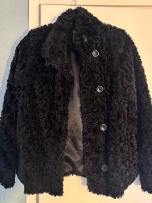 Svart teddyjacka med knappar - Mysig svart kort teddyjacka/teddykappa med stora knappar framtill och fluffig struktur. Jackan har klassisk krage och är helfodrad för extra komfort. Den är i barnstorlek 170 men passar någon med storlek XS eller XXS beroende på vilken passform man vill ha. 