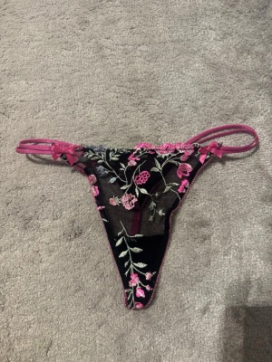 Svart mesh stringtrosa med blommor - Snygg svart stringtrosa i mesh med broderade blommor i rosa, grönt och lite blått. Trosan har dubbla tunna sidoband och fina små rosetter på sidorna. Perfekt för dig som gillar detaljer och en feminin look.