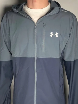 Blå vindbreaker från Under Armour - Snygg tvåfärgad blå vindjacka från Under Armour med huva och dragkedja framtill. Jackan har en bröstficka med dragkedja och logga på vänster sida. Kom med bud, pris är ej huggit i sten!🤩