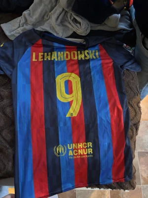 Barcelona matchtröja Lewandowski #9 - Säljer en FC Barcelona matchtröja med Lewandowski och nummer 9 på ryggen. Tröjan har klassiska blå och röda vertikala ränder, gula detaljer och sponsorloggor från Spotify och UNHCR. Tillverkad i lätt och ventilerande material, perfekt för fotbollsträning.