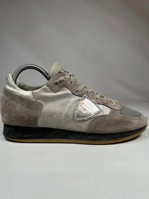 Grå sneakers från Philippe Model - Säljer ett par grå sneakers från Philippe Model med detaljer i mocka och mesh. Skorna har silverfärgade inslag och klassisk snörning. Sidan pryds av den ikoniska sköldloggan och sulan är svart med beige yttersula. Perfekta för en stilren vardagslook.