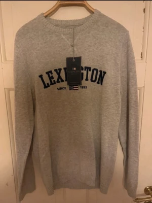 Grå stickad tröja från Lexington - Snygg grå stickad tröja från Lexington med mörkblå text och flaggdetalj framtill. Rund halsringning och långa ärmar. Perfekt till en avslappnad och stilren look. Materialet känns mjukt och bekvämt.