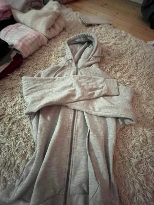 Ljusgrå hoodie från 157 med dragkedja - Ljusgrå hoodie från 157 med dragkedja framtill och klassisk huva. Tillverkad i mjukt material som känns skönt mot huden. Perfekt för chill dagar eller när du vill ha en enkel och stilren look. Långa ärmar och ribbade muddar för extra komfort. Passar jättebra och är i storlek S!