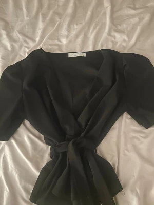 Svart omlottopp från Sisters Point - Svart omlottopp från Sisters Point med knyt i midjan och korta ärmar. Toppen har en v-ringad design och är tillverkad i ett lätt, mjukt tyg som ger en snygg siluett. Perfekt att styla med jeans eller kjol.