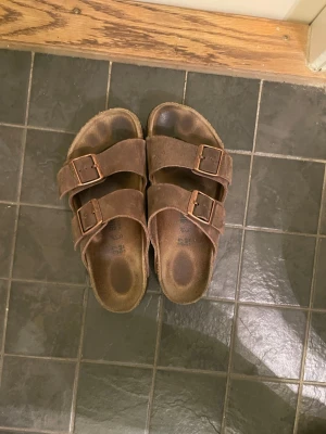 Bruna birkenstocks  - Bruna sandaler från Birkenstock med två breda remmar i skinn och justerbara metallspännen. Sval design med öppen tå och platt sula i kork för extra komfort. Perfekta för sommaren och enkel att matcha med olika outfits.