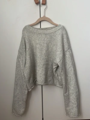 Grå croppad stickad tröja - Mysig croppad stickad tröja i ljusgrått med bred halsringning och långa, vida ärmar. Tröjan har en avslappnad passform och ribbstickade detaljer vid halsen. Perfekt för lager på lager och chill dagar.