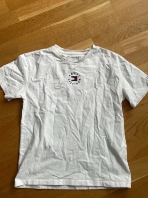 Vit Tommy Hilfiger t-shirt - Säljer en klassisk vit t-shirt från Tommy Hilfiger med rund hals och korta ärmar. På bröstet finns ett broderat Tommy Hilfiger-logo i blått, rött och vitt. Tillverkad i mjuk bomull som känns skön mot huden. Perfekt till jeans eller shorts.