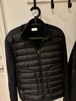 Svart Moncler Cardigan med dragkedja - Svart Cardigan från Moncler med quiltad framsida och stickade ärmar. Jackan har hög krage, två sidofickor med dragkedja och Moncler-logga på vänster ärm. Snygg, stilren och perfekt för kyliga dagar. Moncler etiketten har lossnat på en sida. Ny pris cirka 10.000, säljs billigare pga av etiketten. Använd cirka 3 gånger. NFC tagg i ärm och i DIST etikett. Kvitto finns.