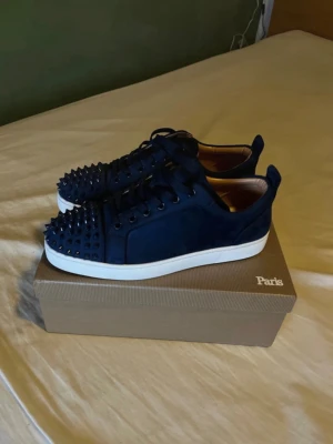 Mörkblå sneakers med nitar. Skorna kommer in 🔜  - Snygga mörkblå sneakers med svarta nitar på tån och ovandelen. Skorna har vit sula och röd undersida, samt snörning. Tillverkade i mocka för en lyxig känsla och edgy look.