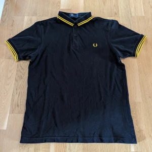 Fred Perry Pike Medium - Säljer en svart och gul Fred Perry pike i storlek M. Fint skick. Står att det är slim fit, men jag skulle säga att den är likadan som den vanliga modellen. 