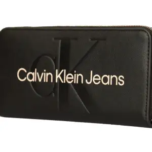 Begagnat skick  Plånbok sculpted zip around svart.   En längre plånbok med logo frambåde tryckt och präglat, plats för 8 kort samt sedlar och mynt.  Mått:19 x 9,5 x 2 cm