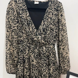 Klänning - Klänning med v-ringning i omlott, elastisk midja och kjol med volanger. Långa ärmar med elastiska ärmslut. Gjord i tunn, vävd kvalitet med foder i trikå.  Ny utan prislapp  