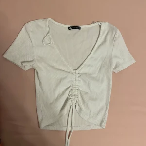 Vit ribbad topp från Zara - En vit ribbad topp från Zara med korta ärmar och en v-ringad hals. Toppen har en snörning framtill som ger en rynkad effekt. Skriv gärna om ni har frågor 💕