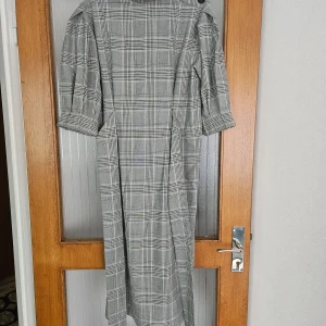 Grå rutig klänning med knappar - En elegant grå rutig klänning med trekvartsärmar och hög krage. Klänningen har knappar på axeln och ett knytband i midjan för en justerbar passform. Den är knälång och har en klassisk design som passar för både vardag och fest. Använd vid ett tillfälle, som ny