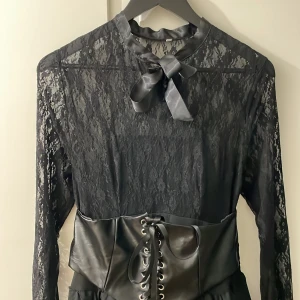 Klänning med tyll o korsett - Svart klänning med spets upptill och snörning i hals, skärp fram som en korsett och bred resår bak i ryggen. Nedtill har klänningen tyll under tyget för att ge mer volym. Oanvänd