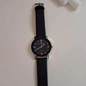Svart klocka från Skylange - En elegant svart klocka från Skylange med en rund urtavla och tydliga vita siffror. Klockan har en svart rem och en silverfärgad spänne. Den är av quartz-typ och har en minimalistisk design.