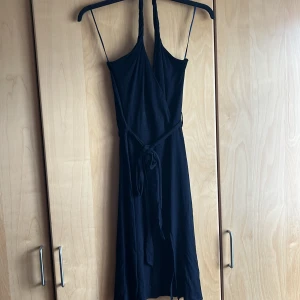 Svart stretchig klänning med halterneck, skärp i midjan och liten slits på sidan - Stretchig halterneck klänning med bar rygg, skärp i midjan och liten slits på sidan. En gång använt, väldigt skön om man gillar att dansa.