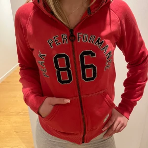 Röd zip-hoodie - Säljer min röda zip-hoodie från Peak Perfomance med supersnygg text som inte kommer till användning 💗den ger en superfin form och är supermysig. (Den har en liten fläck vid texten men syns knappt, skriv till mig för bild)