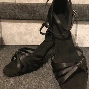 Dansskor oanvända storlek 38 svart satin - 24,8 cm innermått, dansskor inköpta dansaffär Danmark. Klackhöjd 7,5 cm. Går reglera mått spännenöver vrist/ankel. Nya/oanvända p g a fotskada. Kan skickas spårbart 
