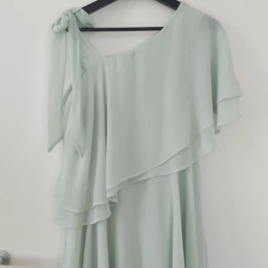 Mintgrön klänning  - En mintgrön kort klänning med volanger och knyt band på ena axeln. Använd endast en gång och är i nyttskick