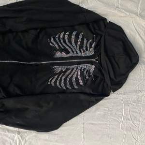 En svart zip up hoodie med rhinestoneskelett, det saknas några stenar på högersidan (se bild 2). Men annars är tröjan i bra skick.