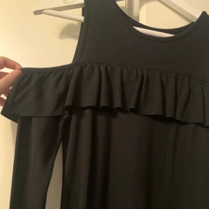 Svart klänning, tonåring  - Svart klänning med ”cut outs” vid axlarna. Volang över bröstet som går hela vägen runt. Har använts sparsamt och köpt på Kappahl. Strlk, 164, passar en 14/15 åring eller någon med storlek xs.