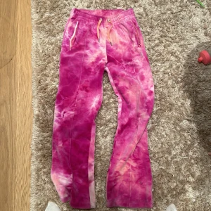 Rosa mjukisbyxor från Juicy Couture - Säljer ett par supermysiga rosa mjukisbyxor från Juicy Couture i storlek XS. Byxorna har ett snyggt batikmönster och är dekorerade med strassdetaljer som bildar bokstaven 'J' på framsidan och 'Juicy' på baksidan. De har en elastisk midja med snörning för bästa passform. Perfekta för en chill dag hemma eller en avslappnad utflykt!