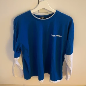 Blå långärmad tröja från Worldwide - Säljer en snygg blå långärmad tröja från Worldwide. Tröjan har vita ärmar som sticker ut under de blåa, vilket ger en cool lager-på-lager effekt. Den är i bra skick och perfekt för både vardag och chill. Passar bra till jeans eller shorts. Skön och stilren design med Worldwide-loggan på bröstet och ryggen.