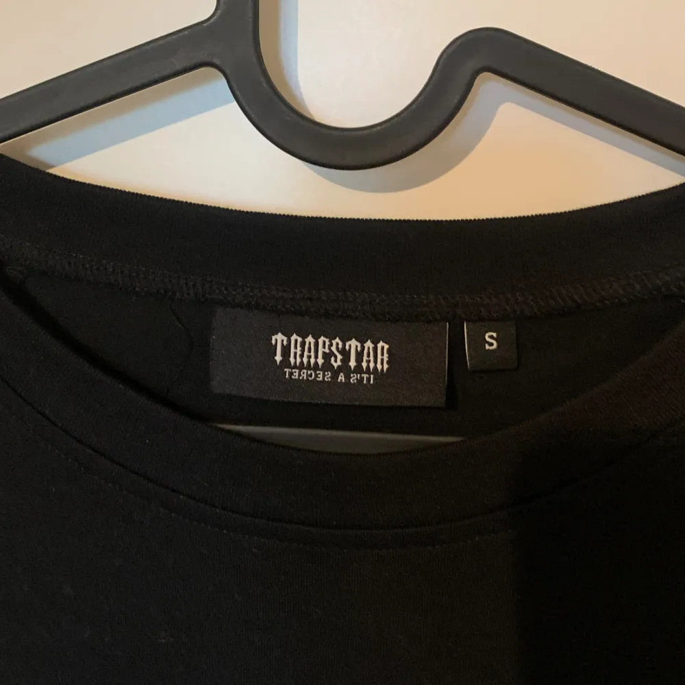 Säljer en svart t-shirt från Trapstar i storlek S. T-shirten har ett coolt tryck på framsidan med texten 'Trapstar' i en gradient från vitt till blått. Den är kortärmad och gjord av bomull, perfekt för vardagsbruk eller fest. Skicket är nyskick, knappt använd.. T-shirts.