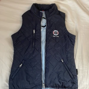 Rid-väst från Covalliero - Säljer en snygg marinblå rid-väst. Västen är i storlek XS och har en dragkedja som man kan öppna upp på ett smart sätt nedtill som ni kan se på bild 4, samt två fickor med dragkedjor. Den har en broderad logga på bröstet med texten 'sinc