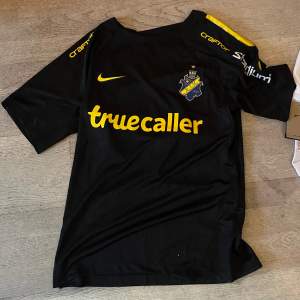 Säljer en svart AIK fotbollströja från Nike med gula detaljer och sponsorer som Truecaller och Craft. Tröjan är i nyskick och har korta ärmar. Perfekt för alla AIK-fans! 💛🖤