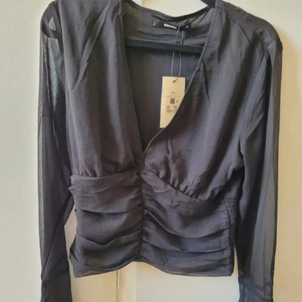 Säljer en elegant svart topp från Bikbok. Den har en v-ringad hals och långa, genomskinliga ärmar. Toppen är åtsittande med rynkade detaljer på framsidan som ger en smickrande passform. Perfekt för både vardag och fest!. Yläosat.