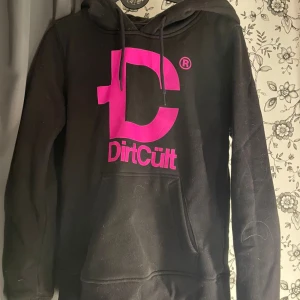 Svart hoodie från DirtCult - Säljer en svart hoodie från DirtCult med en stor rosa logga på framsidan. Den har en stor ficka på magen och en justerbar huva med snören. Perfekt för höst och vinter, supermysig och varm! 💖