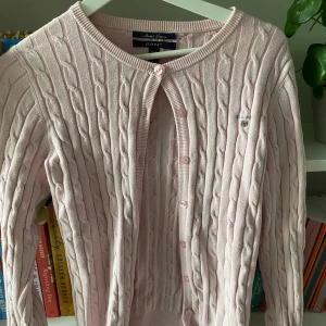 Ljusrosa kofta från GANT - Säljer en superfin ljusrosa kofta från gant. Den är gjord av stretchig bomull och har ett snyggt kabelstickat mönster. Koftan har långa ärmar och knäpps med små rosa knappar framtill. Perfekt för både vardag och fest! 💖