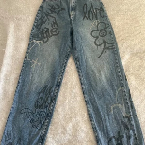 Blå jeans med graffiti-mönster - Ett par blå jeans med graffiti-mönster i svart och grått. Jeansen har en avslappnad passform och är dekorerade med olika ord och teckningar. De har två bakfickor och en hög midja.