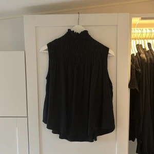 Svart ärmlös topp - Säljer en snygg svart ärmlös topp. Den har en hög krage och är gjord av ett lätt och luftigt material. Perfekt för både vardag och fest!