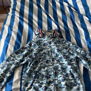Camouflage hoodie från BAPE - Säljer en snygg camouflage hoodie från BAPE. Den har ett coolt hajmotiv på huvan och broderade bokstäver 'WGM' i rött, blått och orange. Hoodien är i blå och grå toner med dragkedja framtill. Perfekt för streetwear-stilen!