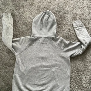 Grå hoodie från GANT - Säljer en snygg och bekväm grå hoodie från GANT. Den har en stor ficka på magen och ett coolt emblem med amerikanska flaggan på bröstet. Perfekt för höst och vinter, och den är nästan oanvänd 🧥✨