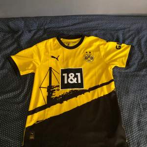 Säljer en gul Borussia Dortmund fotbollströja från Puma. Tröjan har korta ärmar och är i nyskick. Den har svarta detaljer och ett tryck med '1&1' på framsidan samt 'DORTMUND' på baksidan. Perfekt för alla BVB-fans!