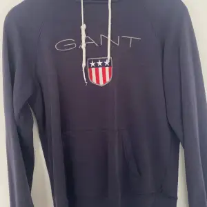 En mörkblå hoodie från GANT med en känguruficka och ett tryck med texten 'GANT' samt en sköld med amerikanska flaggan på framsidan. Hoodien har en justerbar huva med vita dragsnören.