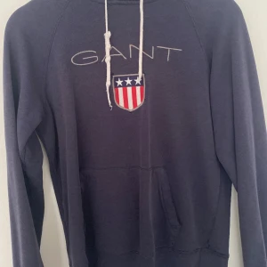 Mörkblå hoodie från GANT - En mörkblå hoodie från GANT med en känguruficka och ett tryck med texten 'GANT' samt en sköld med amerikanska flaggan på framsidan. Hoodien har en justerbar huva med vita dragsnören.