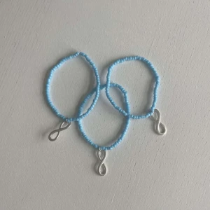 Egengjort pärlarmband  - Tre egengjorda pärlarmband med blåa pärlor. Armbanden har en varsin berlock i silver som avbildar ett evighets tecken. 