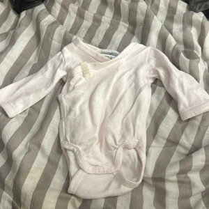 Body  - En rosa body för bebisar i storlek 50, den är ljus rosa med vita linjer på, den har inga fläckar och är inte trasig nånstans 