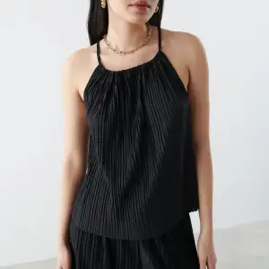 Oanvänd. Denna tröja är från Bianca Ingrosso kollektion med Gina tricot, modellen är ”Tamara singlet”. Plisserat linne med tunna axelband som korsas i ryggen. Linnet är svart och har rynkade detaljer vid halsringningen. Denna tröja säljs inte längre.