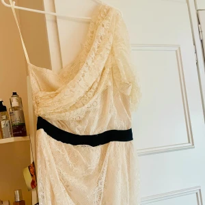 Marlene Birger Design klänning creme spets Student Fest - Vacker spetsklänning i creme färg med svart band i midjan. Design av Marlene Birger.  Dragkedja i sidan. Underklänning i siden. Snyggt fall one shoulder. Storlek S. Passar perfekt till student eller sommarens evenemang och festligheter  Ord pris 1200