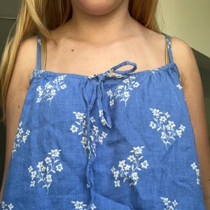 Blått linne med vita blommor  - Super gulligt sommrigt linne med vita fina blommor på. Finns en detalj där man kan knyta en rosett framtil på linnet. Super bra skick och det finns inga tecken på att det har använts. 